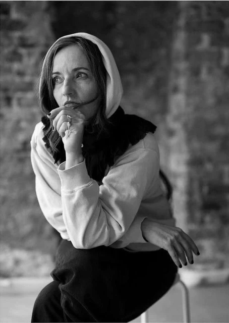 Monochrome portrait of an adult woman in hooded sweatshirt, seated, hand to face, against blurred brick background, thoughtful gaze.