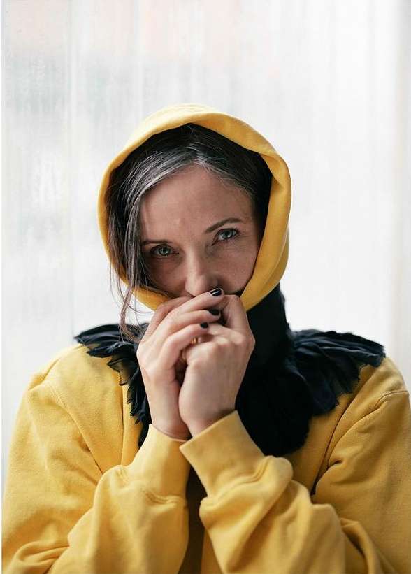 Portrait of an adult woman wearing a yellow hooded sweatshirt with a black ruffled collar, posing in front of a bright window.
