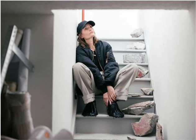 Woman in a bomber jacket and baseball cap sits on concrete stairs with fallen debris, looking up, in an indoor setting with neutral light.
