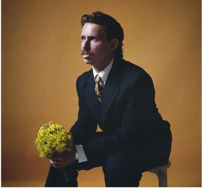 Portrait of a man with a mustache in a dark suit holding yellow flowers, seated against a yellow background, looking away.