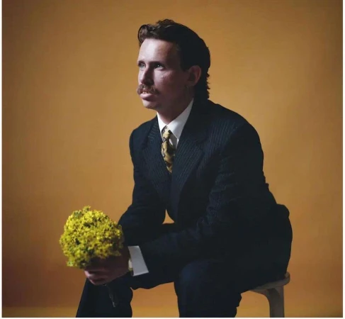 Portrait of a man with a mustache in a dark suit holding yellow flowers, seated against a yellow background, looking away.