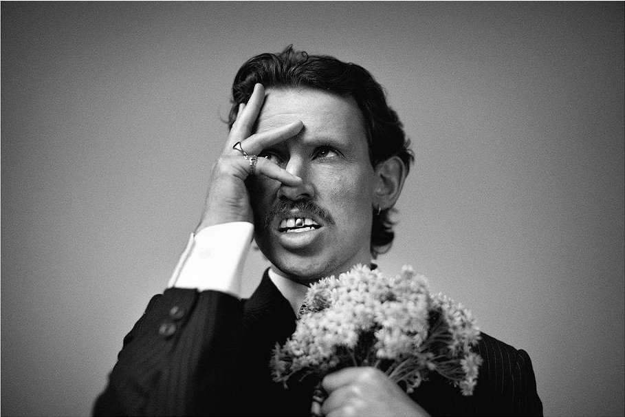 Monochrome portrait of a man in a suit holding flowers, with a hand near his face, set against a simple background.
