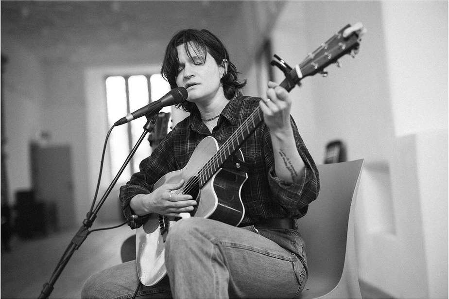 A woman is playing an acoustic guitar while singing into a microphone. She is sitting on a chair, indoors, with natural lighting.
