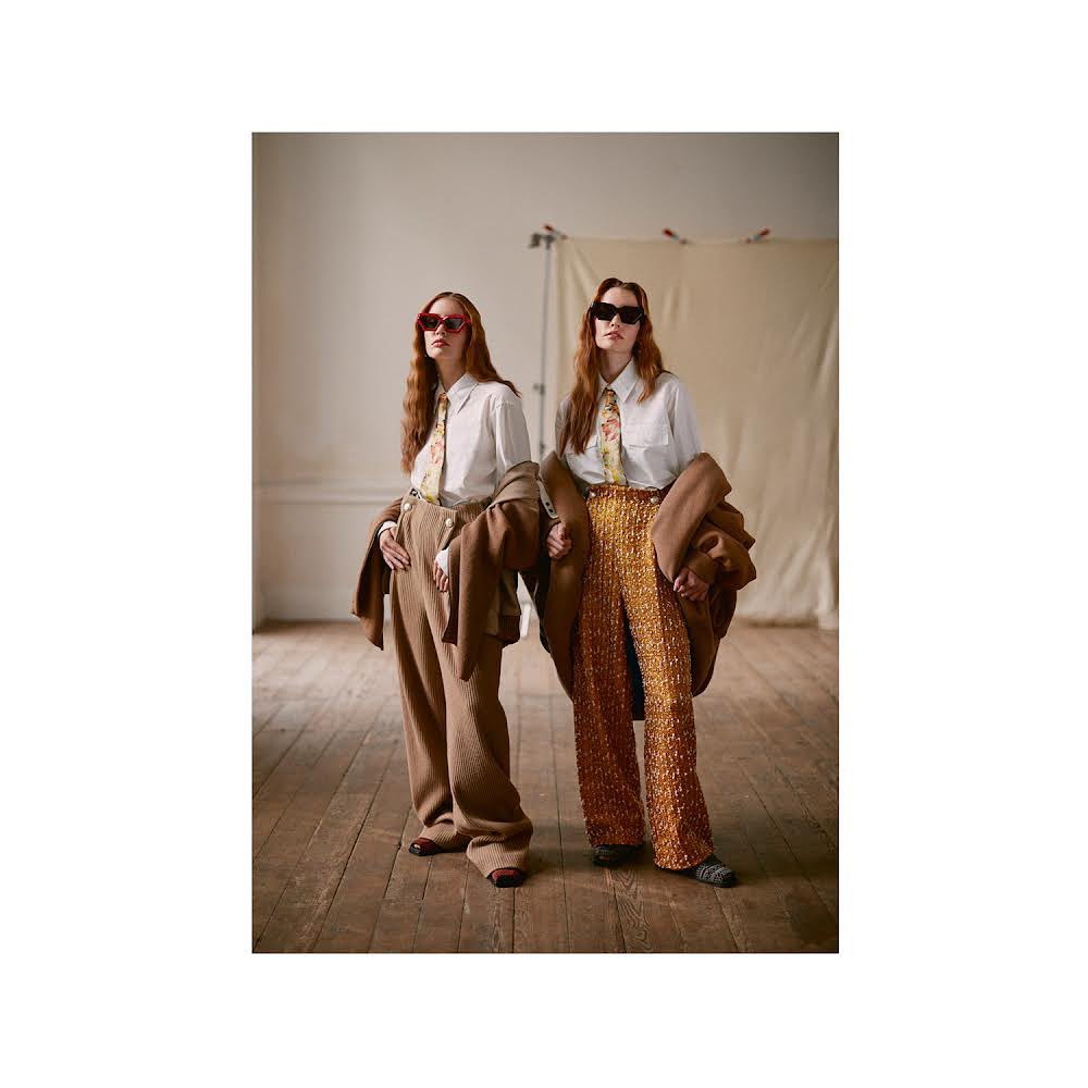 Two women pose in a photo studio wearing matching shirts, ties, sunglasses, and jackets; one in brown pants, the other in orange.