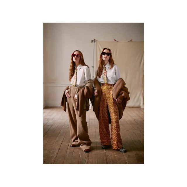 Two women pose in a photo studio wearing matching shirts, ties, sunglasses, and jackets; one in brown pants, the other in orange.