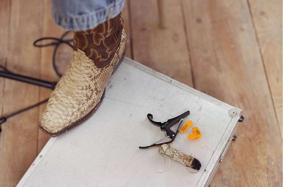 Snakeskin cowboy boot stands on a silver metal case with guitar accessories and a bundle of sage, set on a wood surface.