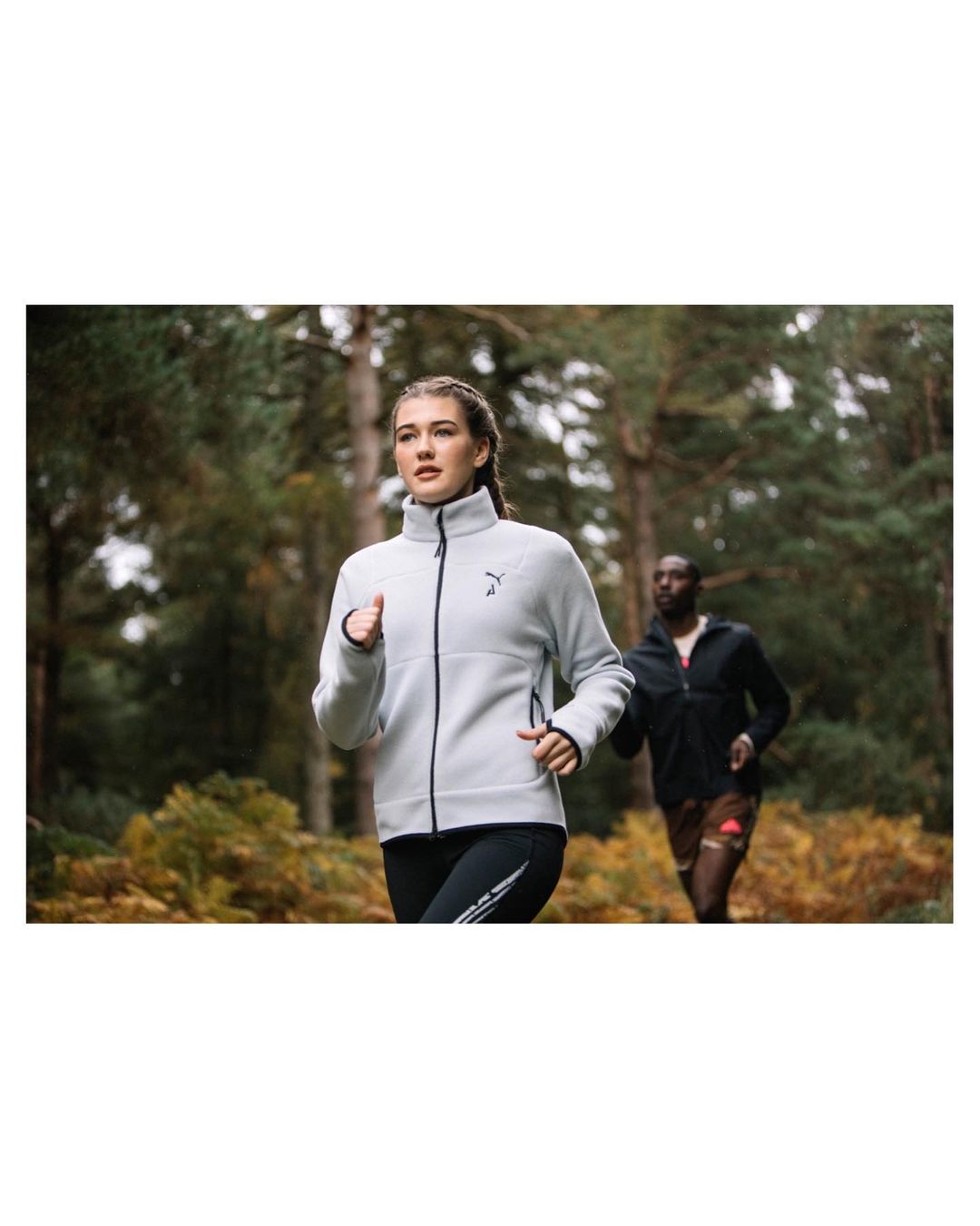 Two runners in athletic wear jog through a wooded area. A woman in a grey jacket and black leggings leads a man in athletic attire.
