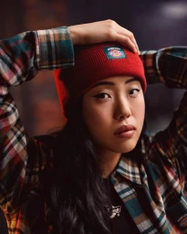 Close-up portrait of a woman wearing a red beanie and plaid shirt with her hands behind her head