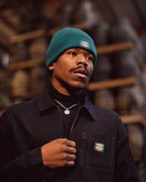 Portrait of a Black man wearing a dark jacket, turtleneck, silver chain necklace, and a green beanie hat.