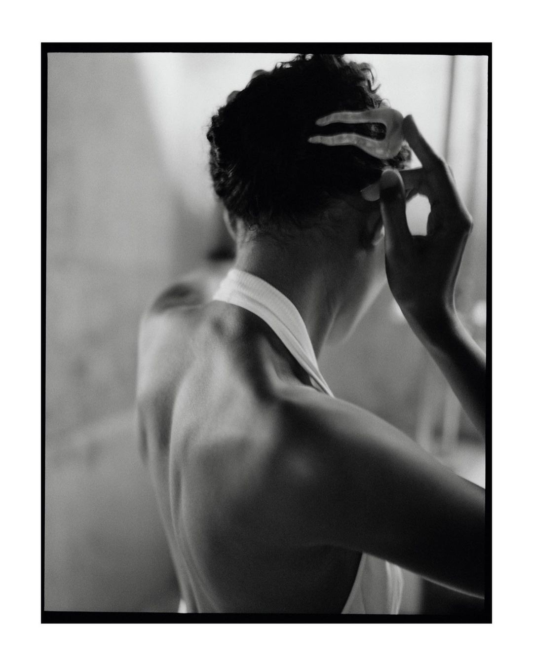 A woman in a white halter top is seen from the back, holding a hair clip in her hand, styled in a black and white photograph.