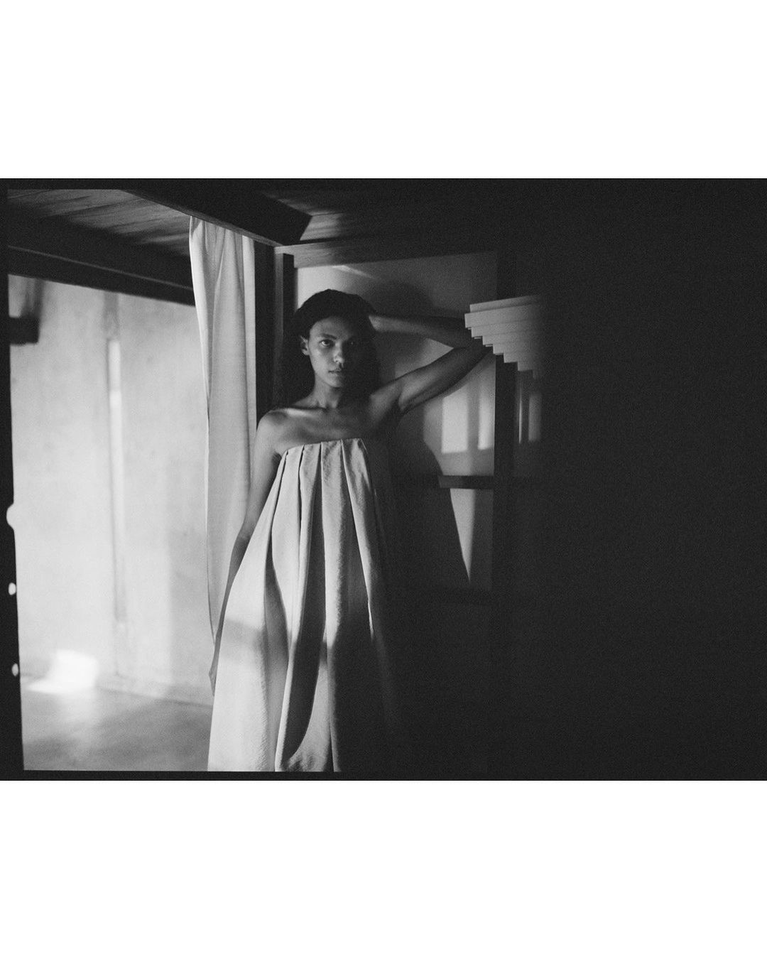 Artistic monochrome portrait of a woman draped in fabric, near a staircase and doorway, with shadows and minimalist decor.