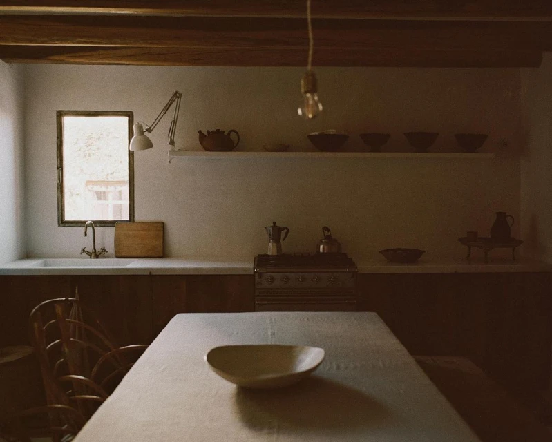 Rustic kitchen interior featuring a table with a ceramic bowl, classic stove, and various pottery pieces arranged on shelves and counters.