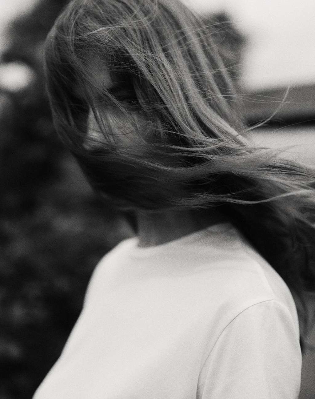 Monochrome portrait of a woman with long, flowing hair, wearing a white t-shirt, with a blurred, natural background, capturing a serene moment.