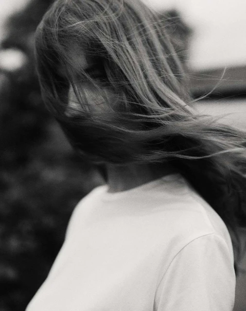 Monochrome portrait of a woman with long, flowing hair, wearing a white t-shirt, with a blurred, natural background, capturing a serene moment.