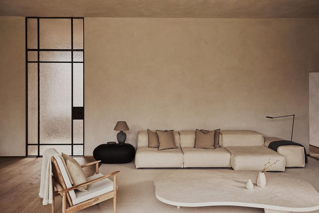 Living room interior featuring a cream sofa, wooden chair, and textured glass door in a minimalist, neutral-toned setting.