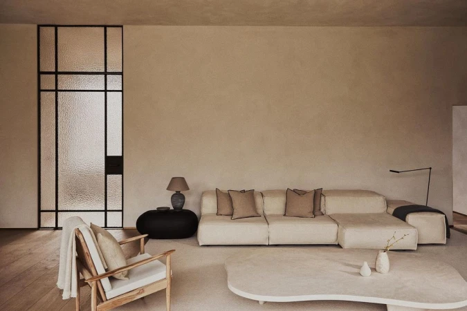 Living room interior featuring a cream sofa, wooden chair, and textured glass door in a minimalist, neutral-toned setting.