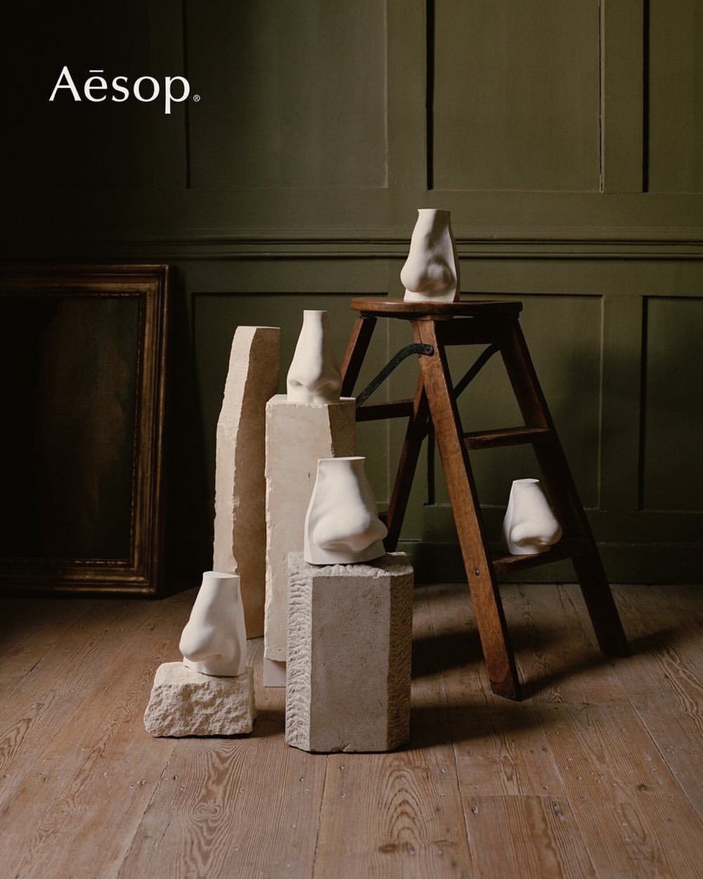 Still life of nose-shaped vases on stone pedestals and wooden ladder against a green wall with an ornate frame and wooden floor.