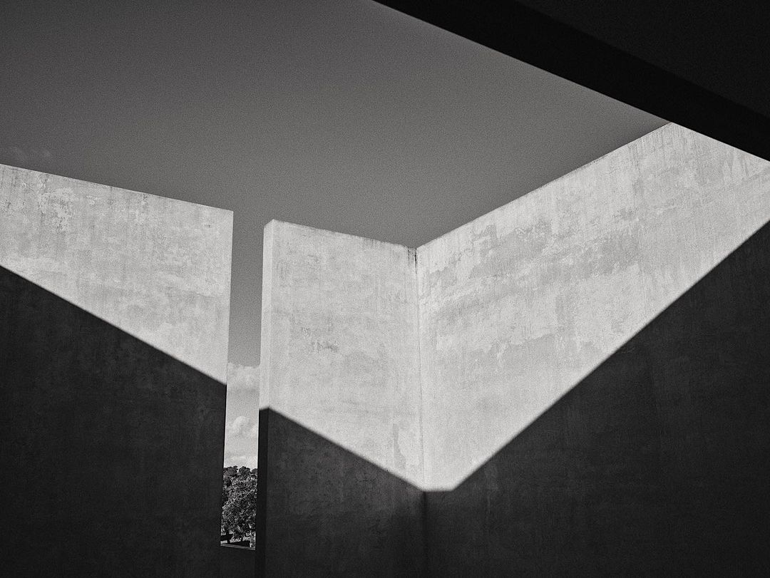 Monochrome architectural shot of concrete walls with angular shadows creating geometric patterns, with a distant view of trees and sky.