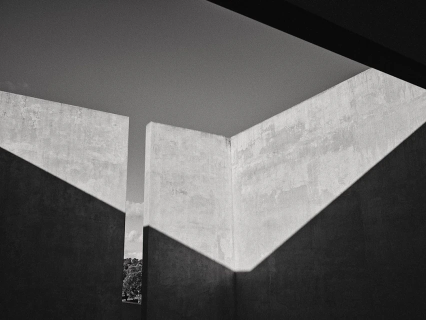 Monochrome architectural shot of concrete walls with angular shadows creating geometric patterns, with a distant view of trees and sky.