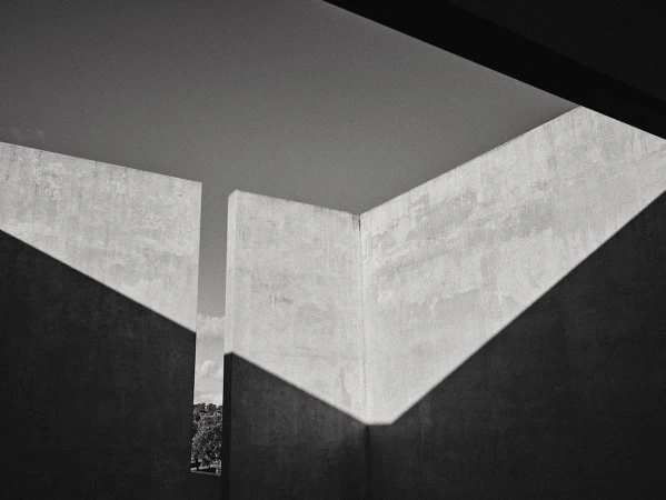 Monochrome architectural shot of concrete walls with angular shadows creating geometric patterns, with a distant view of trees and sky.
