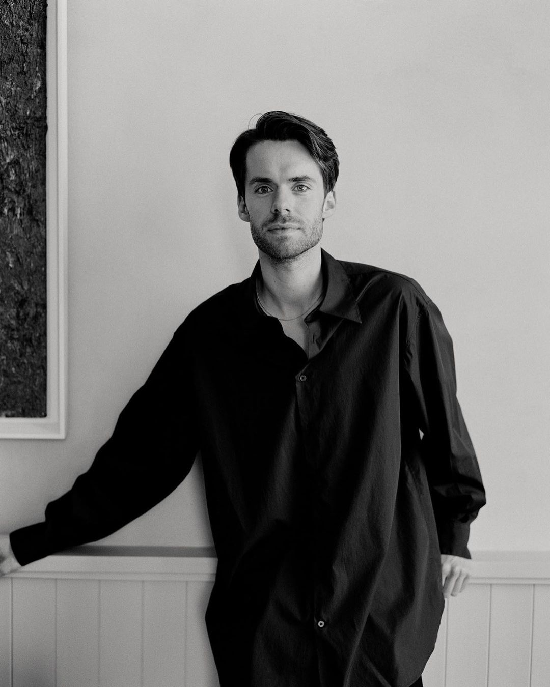 Portrait of a young adult male in a dark, oversized shirt leaning against a white wall in a black and white photo, indoor setting.
