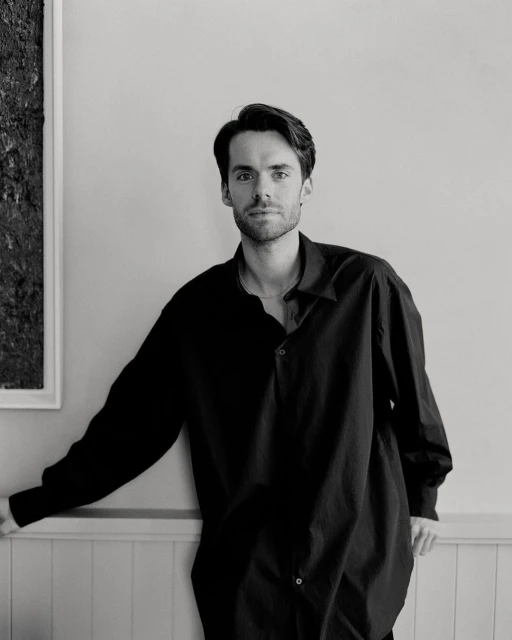 Portrait of a young adult male in a dark, oversized shirt leaning against a white wall in a black and white photo, indoor setting.