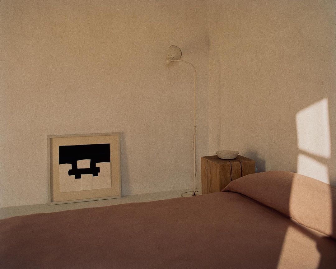 Minimalist bedroom interior featuring a bed with mauve bedding, an artwork, a wooden bedside table, and a floor lamp in soft light.