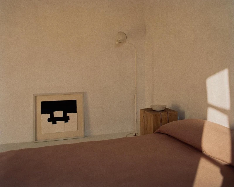 Minimalist bedroom interior featuring a bed with mauve bedding, an artwork, a wooden bedside table, and a floor lamp in soft light.