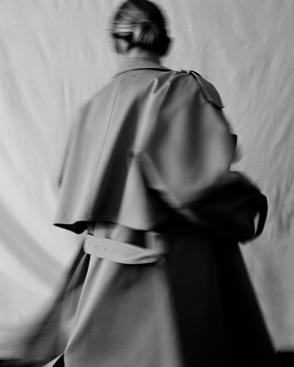 A blurred, monochrome fashion shot of a woman in a tan coat, emphasizing style and motion with a simple background.