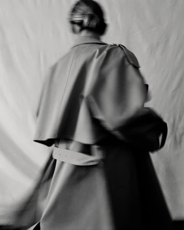 A blurred, monochrome fashion shot of a woman in a tan coat, emphasizing style and motion with a simple background.