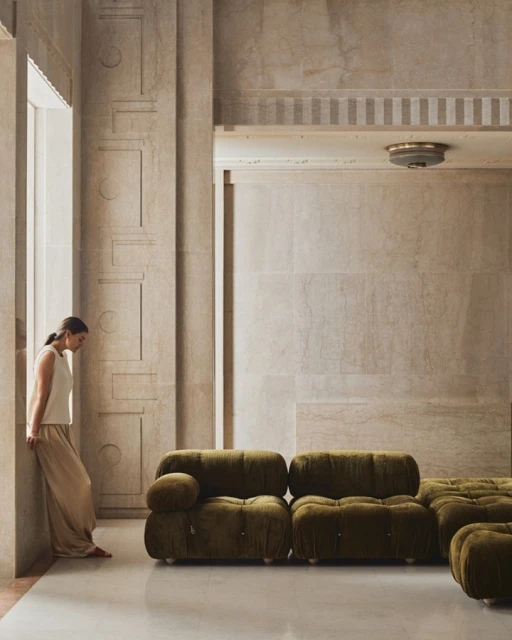 Olive velvet lounge seating in a marble room with a woman leaning against a pillar by a window, casting shadows and natural light.