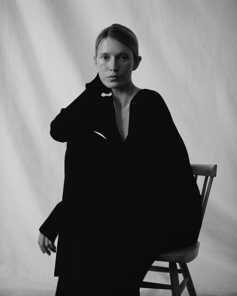 Portrait of a woman wearing a black garment seated on a wooden chair against a draped backdrop, photographed by Rich Stapleton.