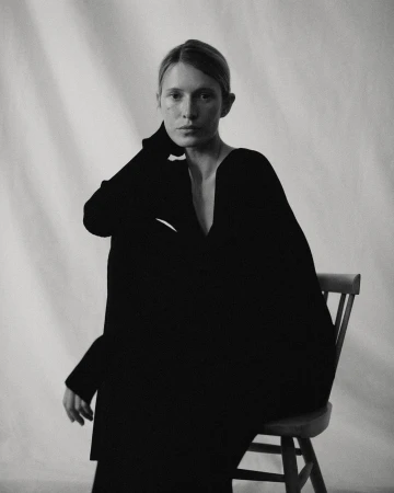 Portrait of a woman wearing a black garment seated on a wooden chair against a draped backdrop, photographed by Rich Stapleton.