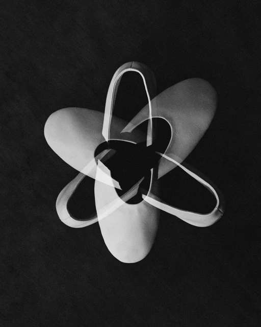 Monochrome image showcasing a flower-like arrangement of three ballet shoes overlapping, presented against a dark background.