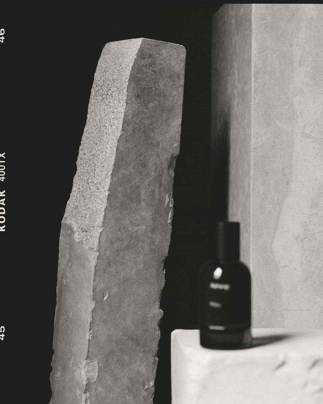 Black and white product photography of a glass perfume bottle on a plinth with rough stone monoliths by Rich Stapleton.