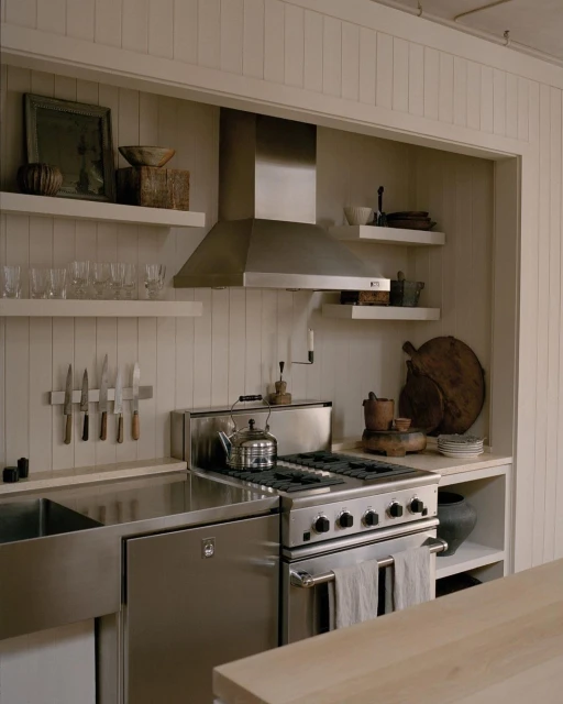Stainless steel stove and sink in a bright, well-lit kitchen with white walls and wooden shelves holding decorative items and utensils.