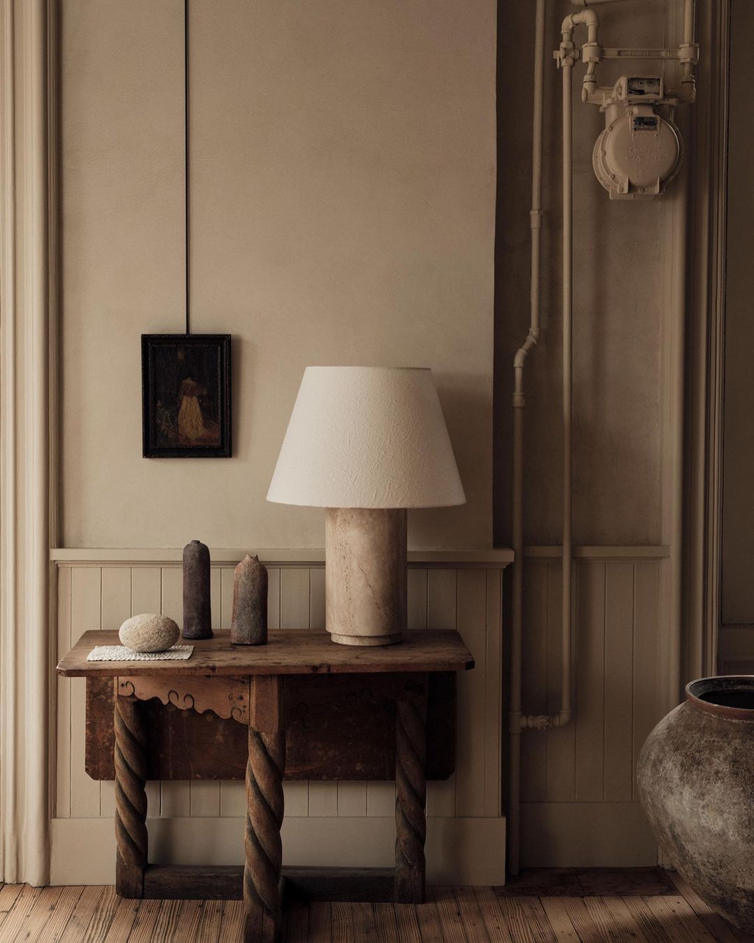 Still life of lamp and decorative objects on wood table in neutral interior with tan walls and wood floor, warm lighting.