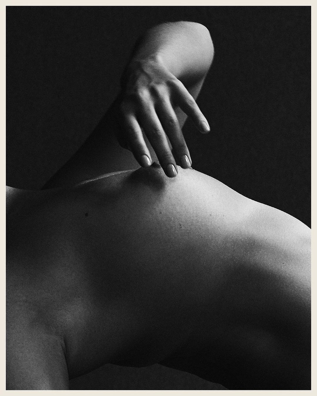 Monochromatic nude photo of an adult, light-skinned human torso, featuring a hand gently touching the body against a dark background.
