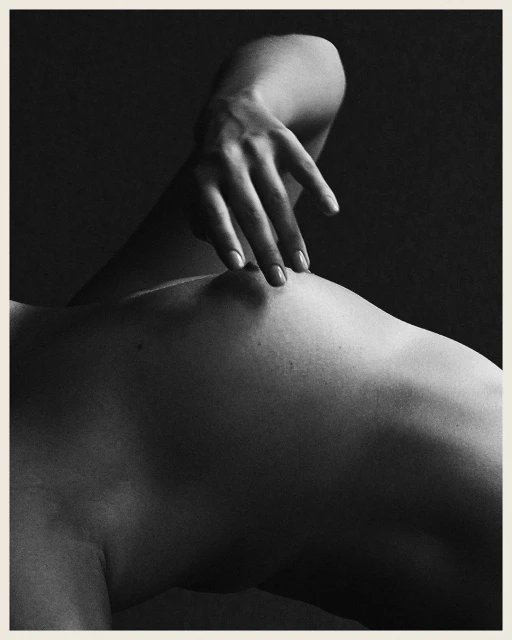 Monochromatic nude photo of an adult, light-skinned human torso, featuring a hand gently touching the body against a dark background.