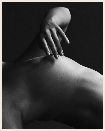 Monochromatic nude photo of an adult, light-skinned human torso, featuring a hand gently touching the body against a dark background.