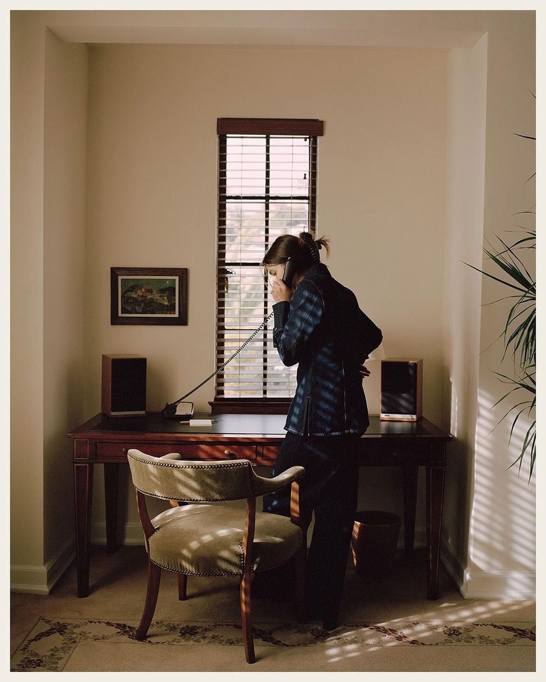 Woman in navy shirt on phone in a sunlit room with a wooden desk, chair, speakers, window with blinds, and houseplant.