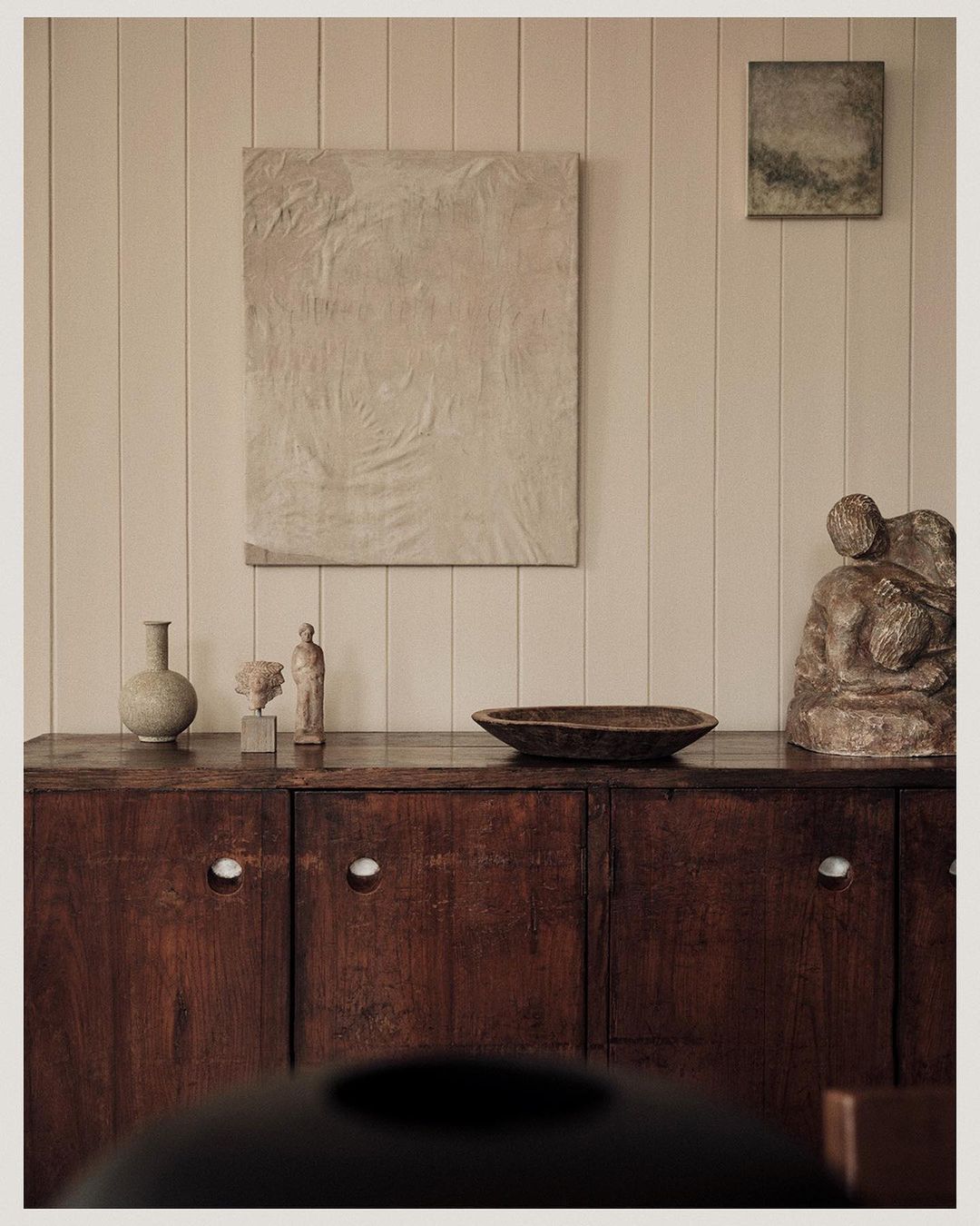 Dark wood sideboard with art, vases, and sculpture in a neutral-toned interior, showcasing home decor and artistic arrangement.