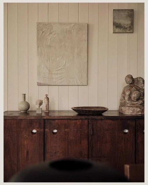 Dark wood sideboard with art, vases, and sculpture in a neutral-toned interior, showcasing home decor and artistic arrangement.