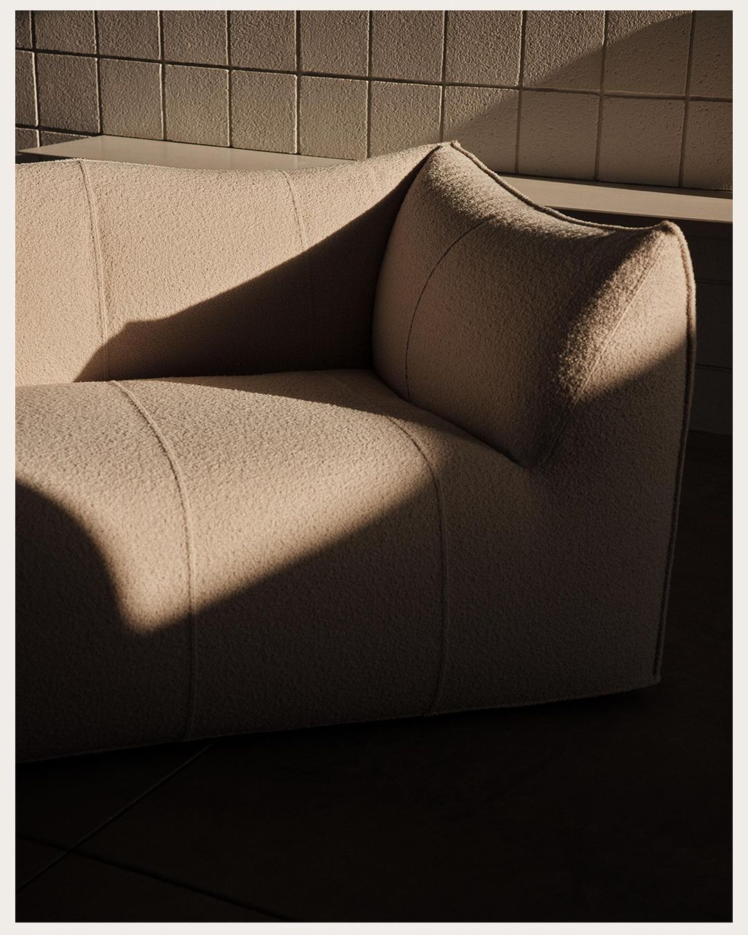 Modern off-white sofa positioned in a room with natural sunlight casting shadows on the furniture and tiled wall in the background.