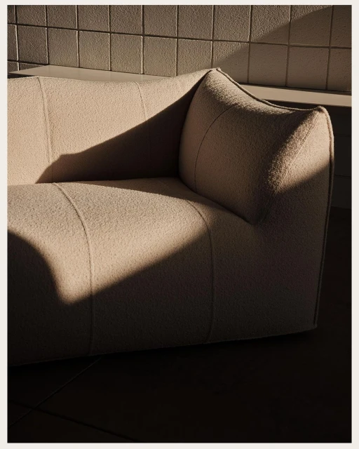 Modern off-white sofa positioned in a room with natural sunlight casting shadows on the furniture and tiled wall in the background.