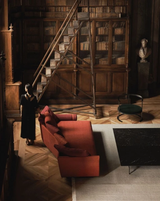 Woman in a black dress stands in a wood-paneled library with a rolling ladder, orange sofa, black table, and herringbone floor.