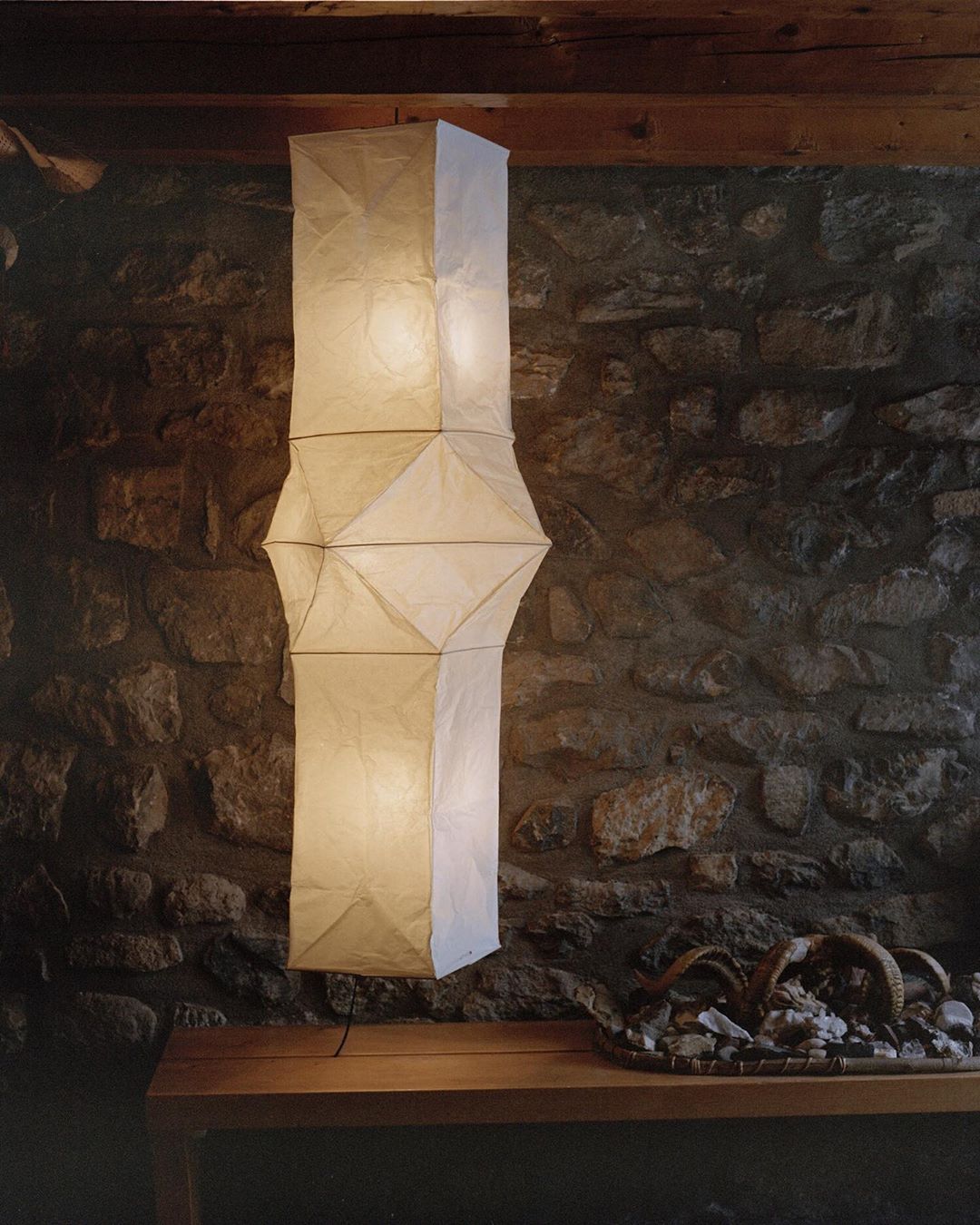 Tall, decorative paper lantern lamp casting light in a rustic room with a stone wall and a wooden table displaying decorative stones.