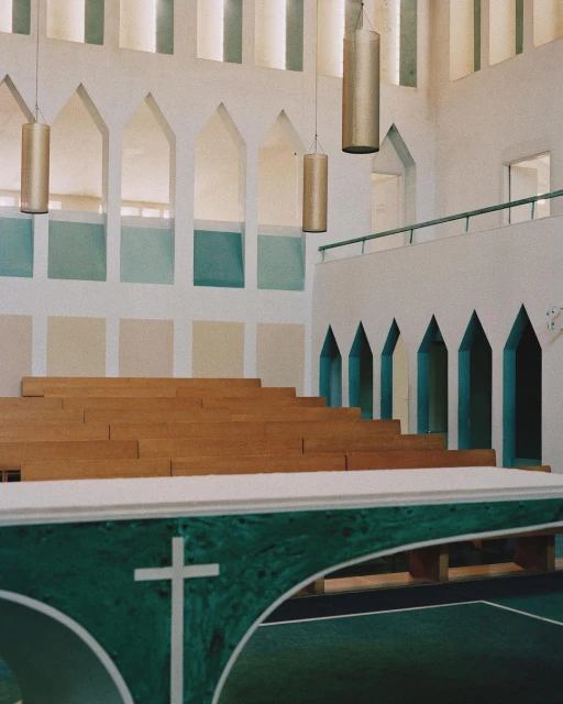 Church interior featuring wooden pews, a green altar with a white cross, and natural light through arched windows, creating a serene atmosphere.