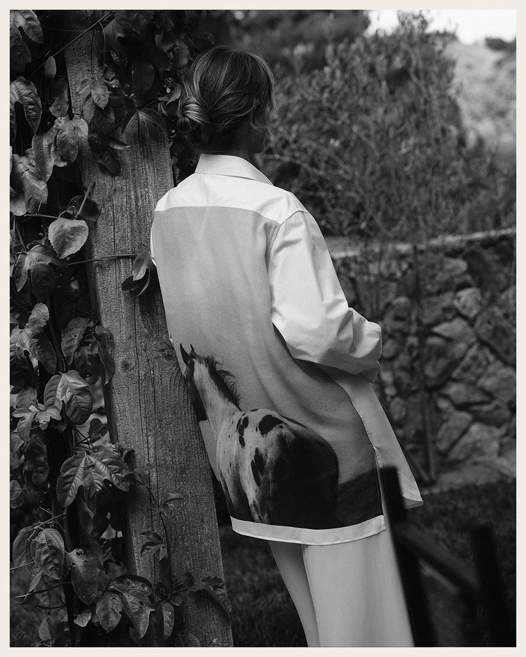 Woman in white shirt with horse print poses against a wooden post outdoors amid trees and plants in a black-and-white fashion shot.