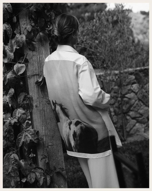Woman in white shirt with horse print poses against a wooden post outdoors amid trees and plants in a black-and-white fashion shot.